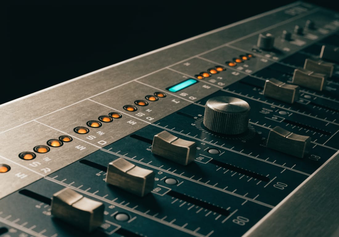 A mixing console channel strip in raking light: brushed aluminium panels, etched scale markings, a row of amber indicator lamps and one narrow cyan segment readout.