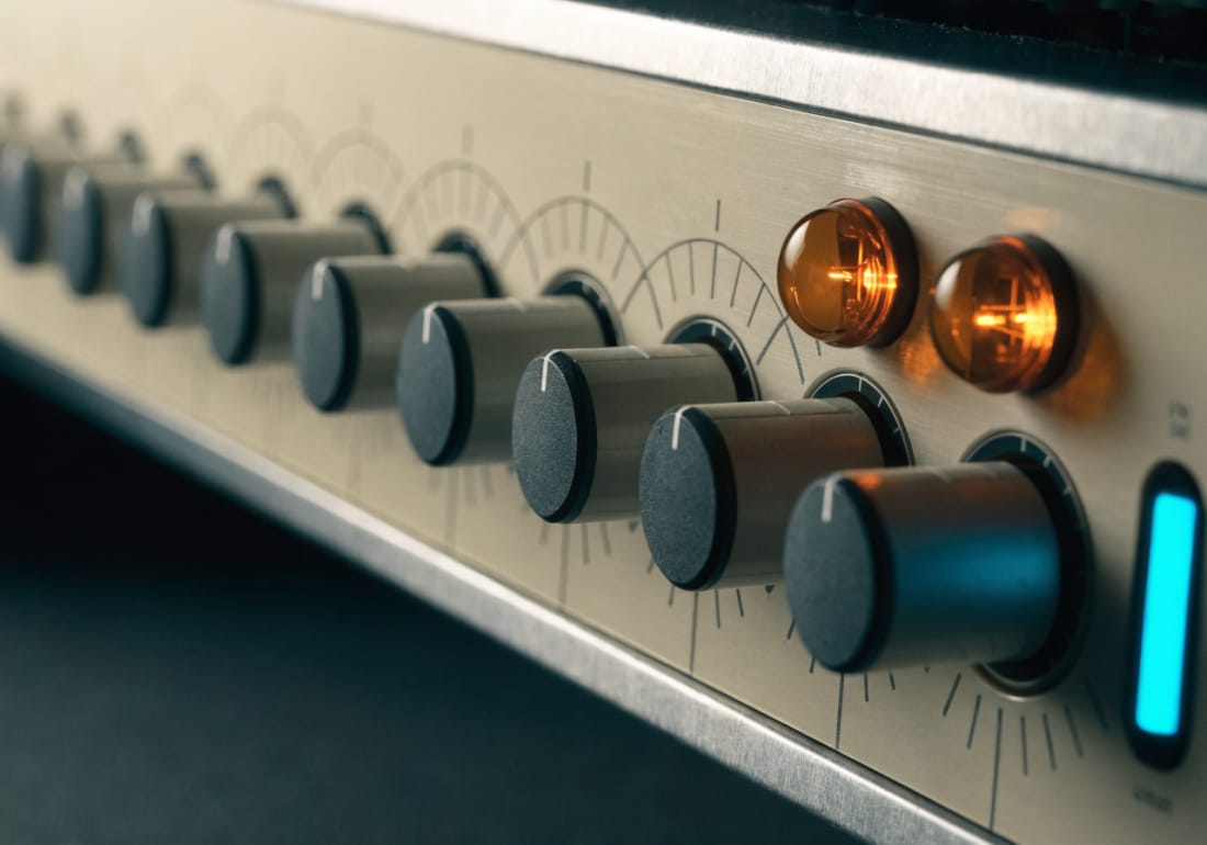 A hardware synthesiser front panel at a shallow angle: a row of metal knobs with white index lines over etched scale arcs, two amber lamps and a cyan indicator.