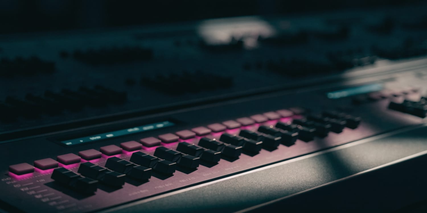 A lighting control desk in a darkened room: rows of faders and square buttons backlit in magenta, with a narrow cyan readout above them.